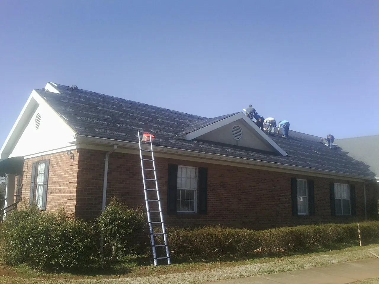 Crew tearing shingles off a brick ranch roof with a ladder set — Emergency Roof Repair work by our crew in Oakfield